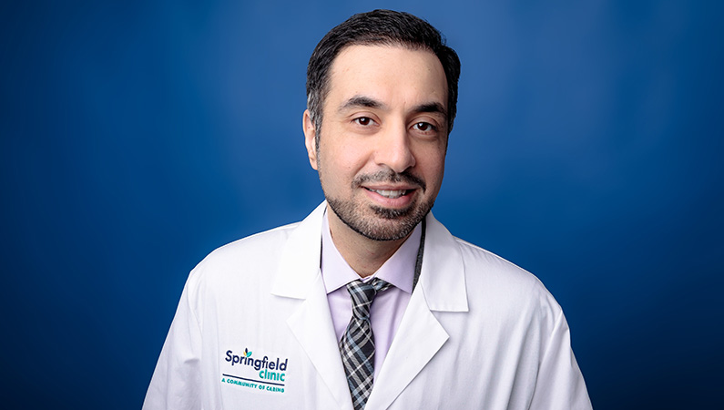 Male doctor in white medical coat smiling in front of navy blue photo backdrop.
