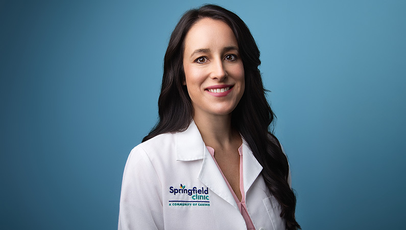 Female smiling wearing white medical coat with blue photo backdrop in background