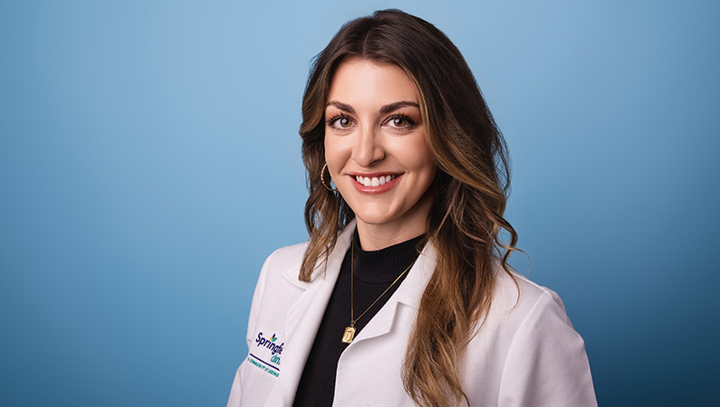 Female nurse with brown hair wearing white medical coat smiling in front of light blue photo backdrop.