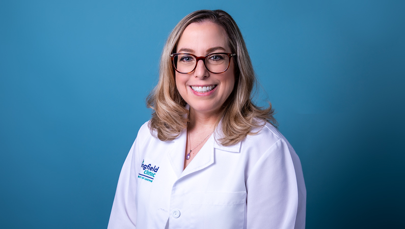 Female with light hair wearing glasses and white medical coat smiling in front of blue background