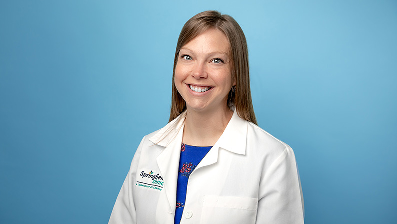 Female nurse in white lab coat smiling in front of light blue backdrop.