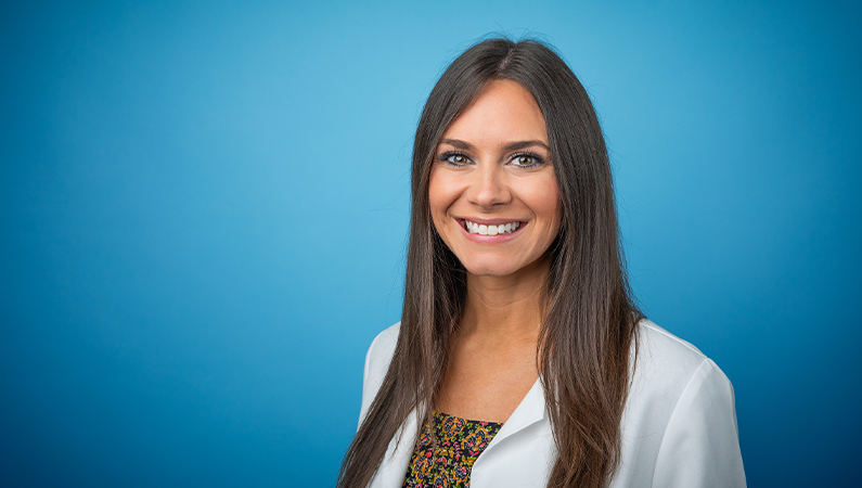 Female nurse practitioner in white coat and long brown hair smiling in front of a light blue backdrop.