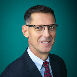 Male family medicine doctor with glasses smiling in front of a teal photo backdrop.