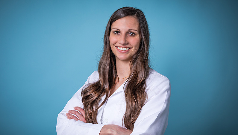 Female nurse practitioner in white coat posing in front of a light blue background.