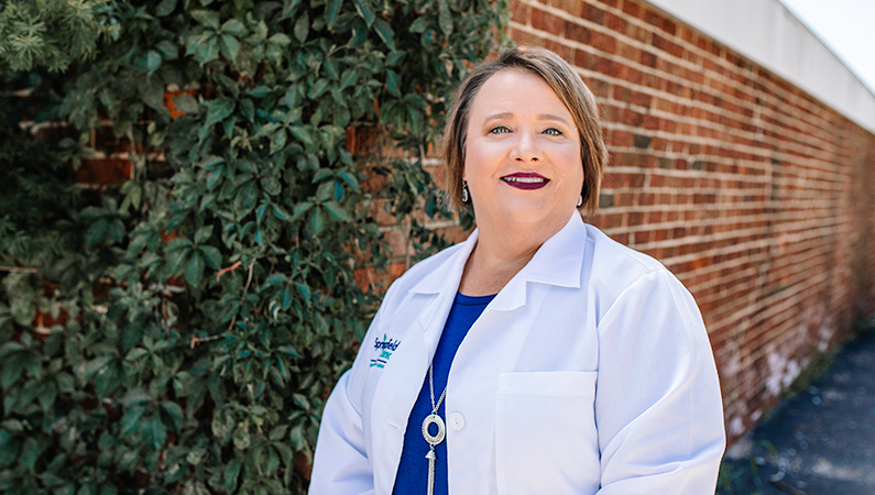 Woman outside smiling in front of a brick wall and green vines.