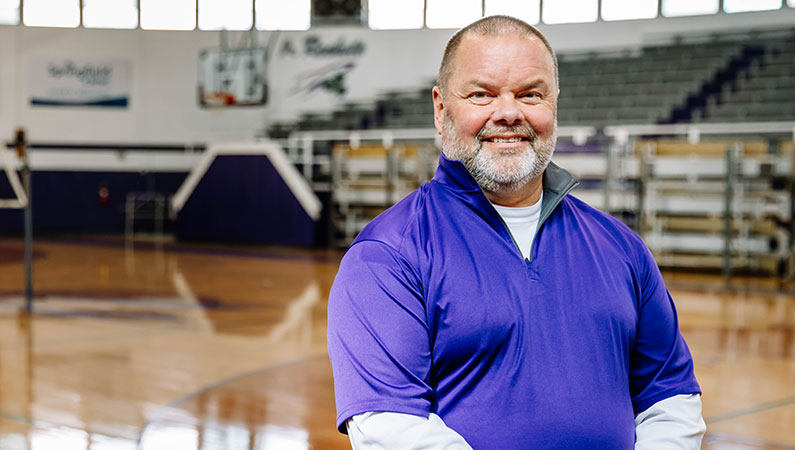 Male athletic trainer wearing purple shirt smiling in school gym setting