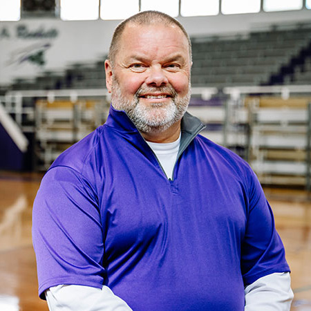 Male athletic trainer wearing purple shirt smiling in school gymnasium.