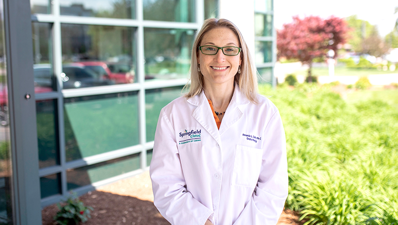 Female physician assistant in white lab coat smiling in front of a glass building.