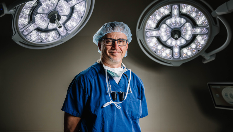 Male surgeon posing in a patient room with surgical lights behind him.