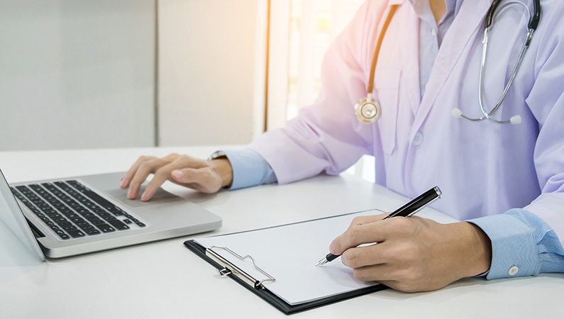 Individual wearing medical coat and stethoscope on computer and writing information down on clipboard