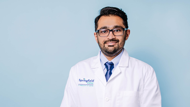 Male doctor wearing a white coat smiling in front of a light blue background.