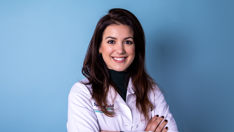 Female with dark hair wearing white medical coat smiling in front of blue background