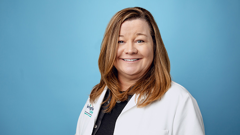 Female nurse practitioner in white medical coat smiling in front of blue photo backdrop.
