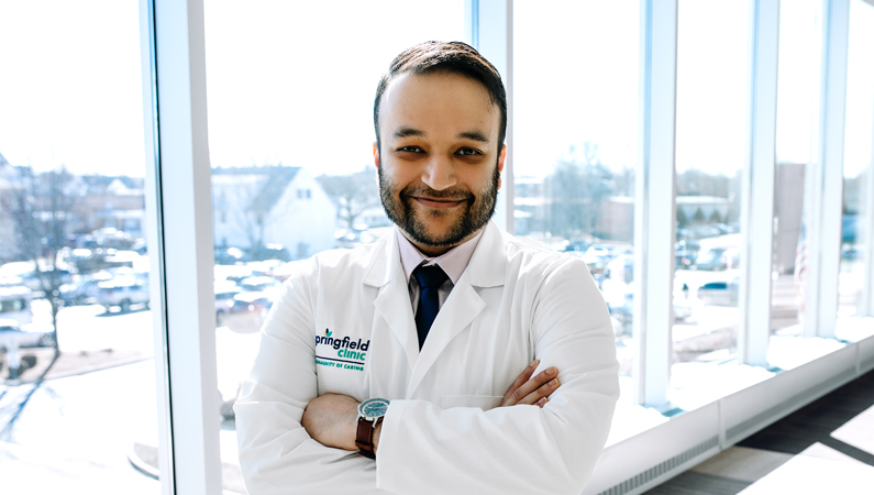 Male wearing white medical coat posing with arms crossed in front of windows