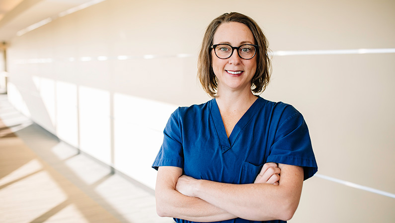 Female medical professional with short brown hair and glasses and arms crossed smiling in a hallway