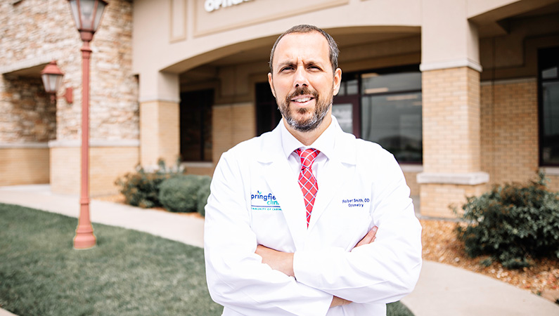 Male wearing white medical coat smiling outside of medical office building