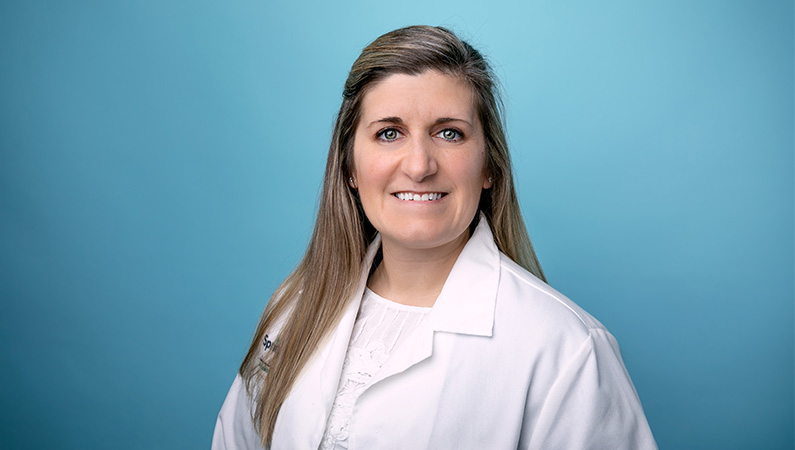 Female nurse practitioner in white medical coat smiling in front of a light blue photo backdrop.