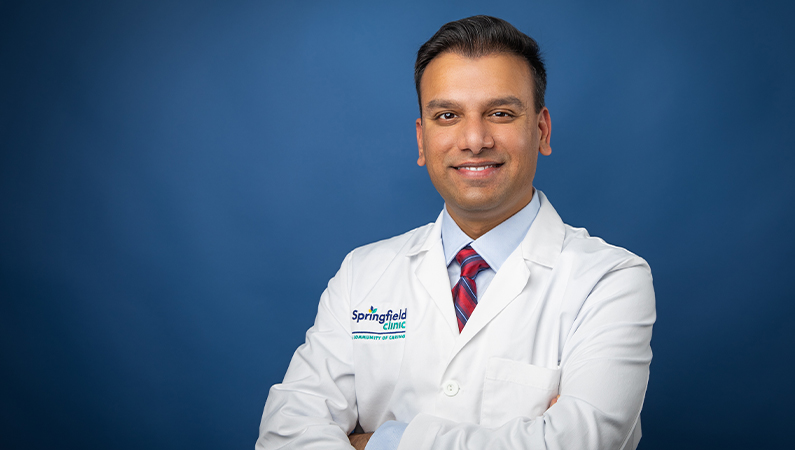 Male hospital medicine doctor in white coat smiling in front of photo backdrop.