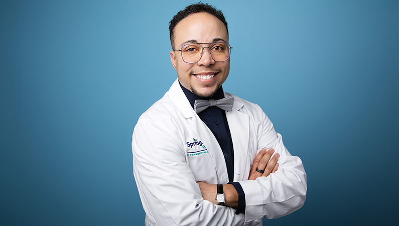 Male radiologist wearing white medical coat smiling in front of light blue photo backdrop.