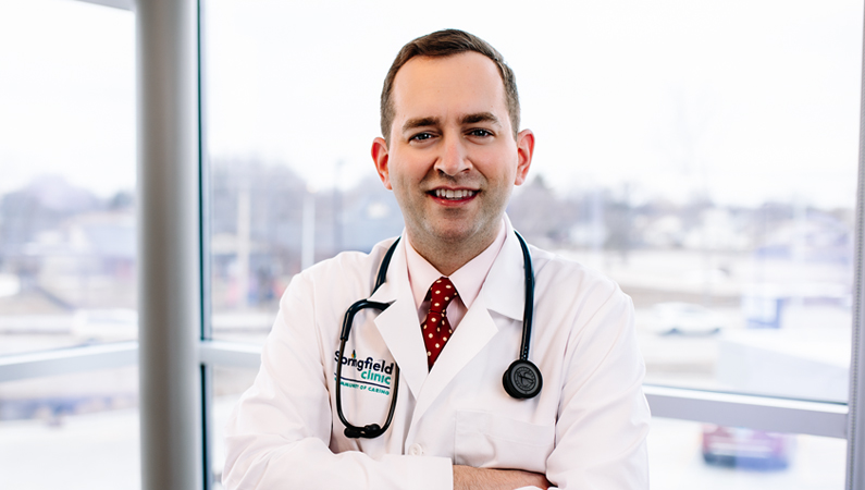 Male wearing white medical coat and stethoscope smiling in front of windows