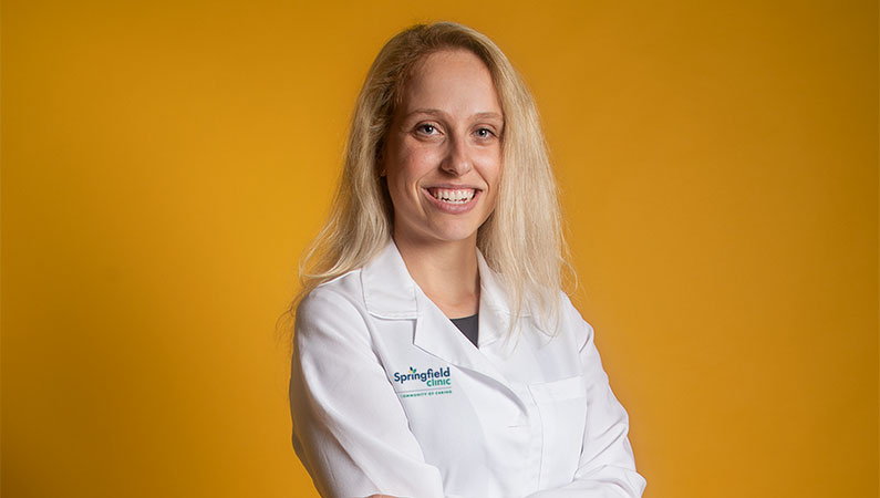 Light haired female medical professional wearing white coat smiling in front of yellow backdrop