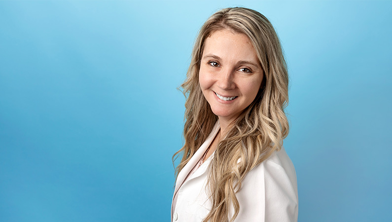 Female nurse practitioner in white lab coat smiling in front of light blue photo backdrop.