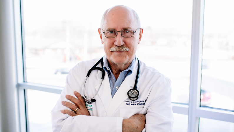 Male wearing glasses and stethoscope with white medical coat with arms folded posing in front of windows