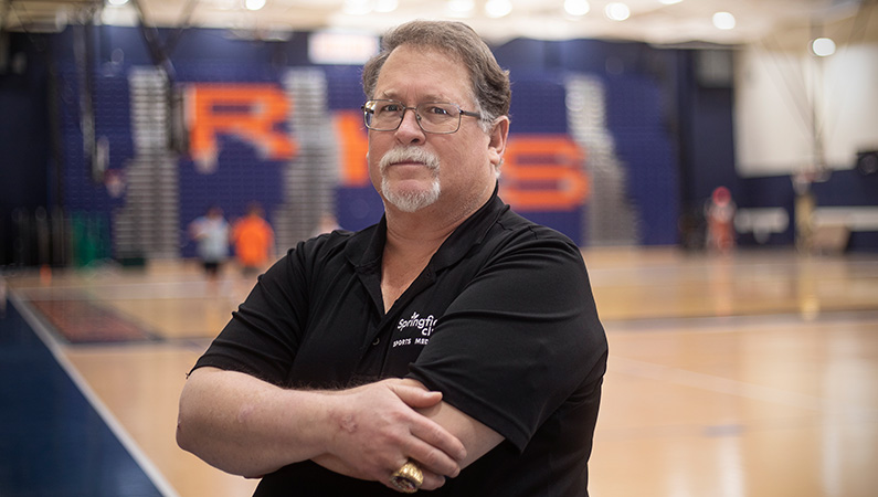 Male wearing glasses with arms folded posing in front of high school gymnasium interior