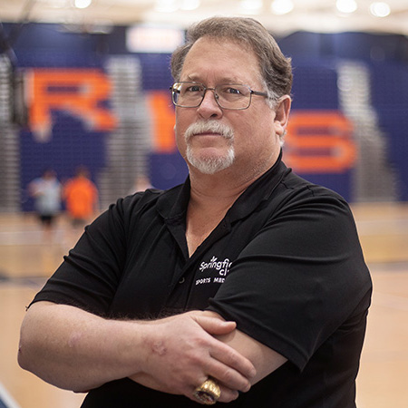 Male athletic trainer smiling with arms crossed in a school gym setting.