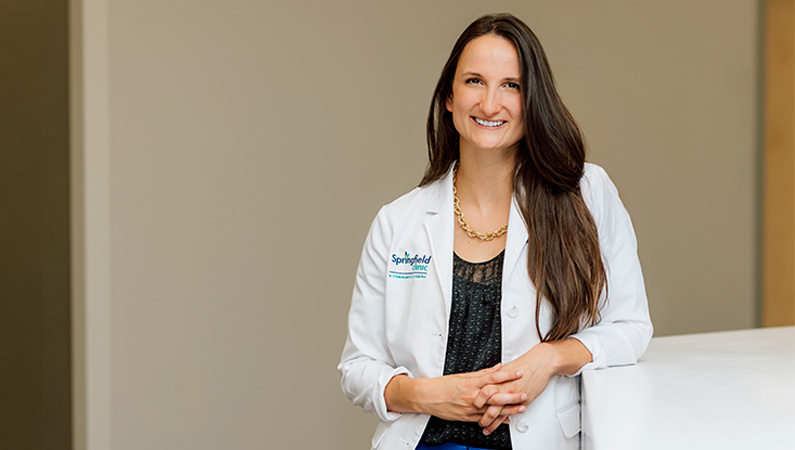 Female doctor in white lab coat smiling in medical facility hallway.