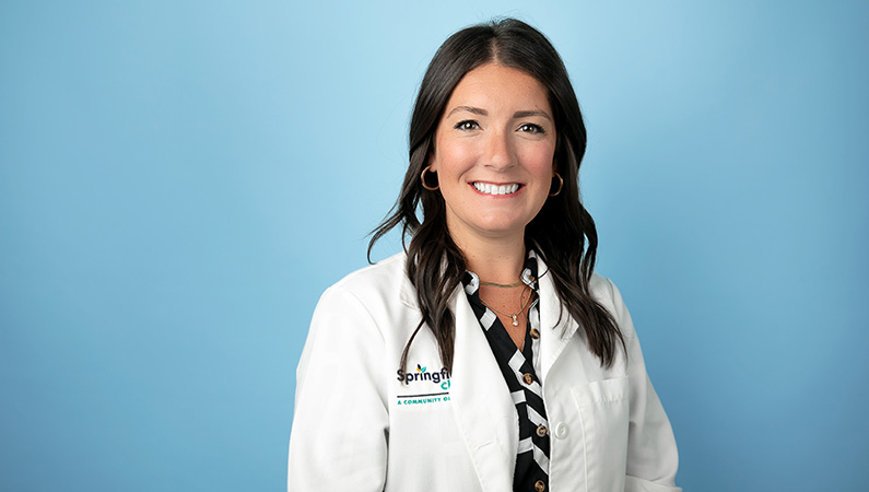 Female nurse practitioner in white medical coat smiling in front of light blue photo backdrop.