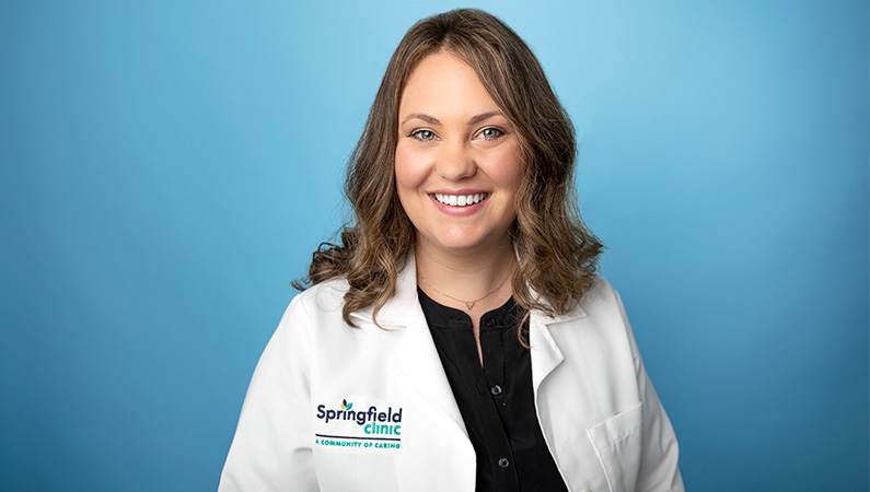 Female nurse in white medical coat smiling in front of light blue photo backdrop.