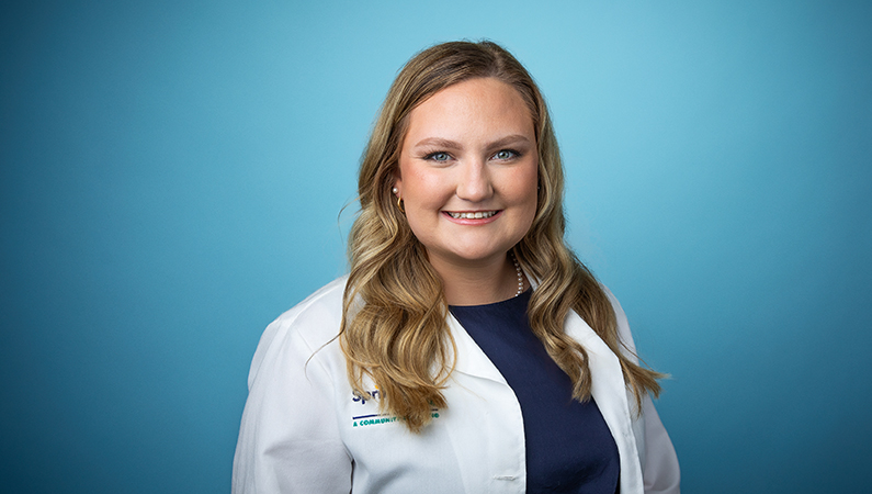 Female wearing white medical coat smiling in front of blue photo backdrop