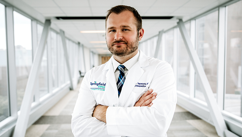 Male with brown hair wearing white medical coat posing with arms folded in hallway
