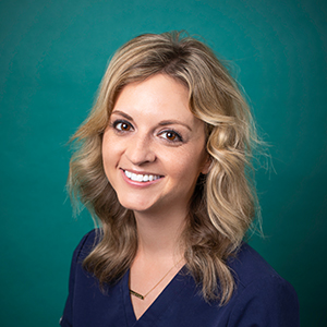 Female dermatologist doctor smiling in front of a teal blue backdrop.