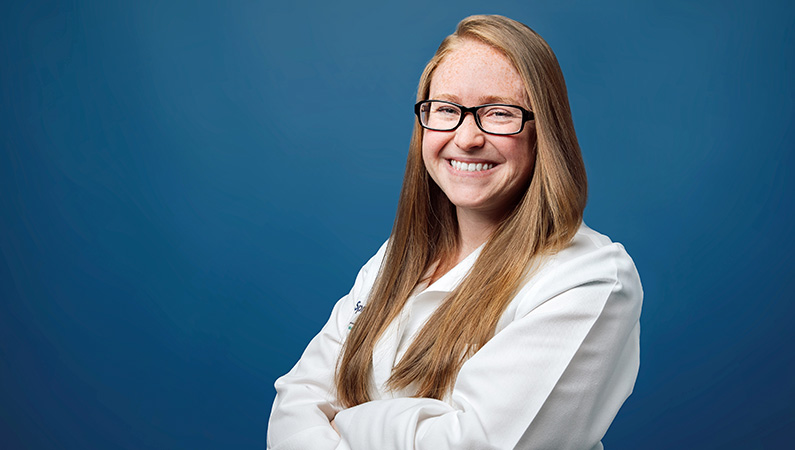 Female hospitalist in white medical coat smiling in front of light blue photo background.