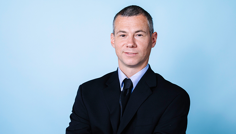 Male nurse practitioner in black suit posing against a blue background.