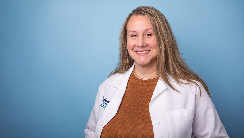 Female nurse practitioner in white lab coat smiling in front of blue photo backdrop.