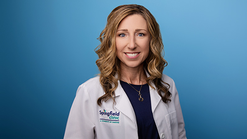 Female nurse practitioner wearing white medical coat smiling in front of light blue photo backdrop.