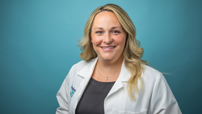 Female nurse practitioner in white coat smiling in front of light blue backdrop.