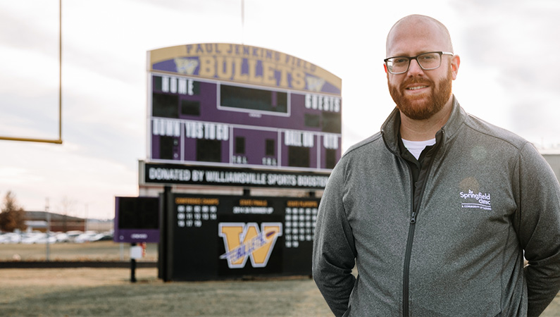 Male wearing glasses and sports zip up smiling in on football field in front of scoreboard