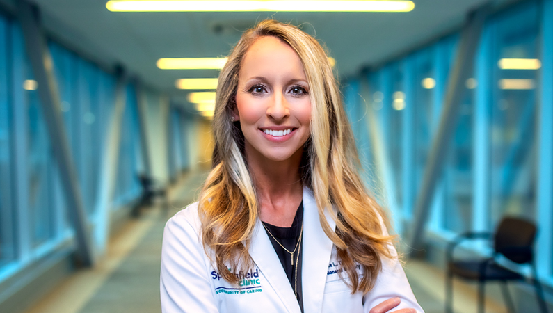 Female with wavy hair wearing a white medical coat smiling in a walkway