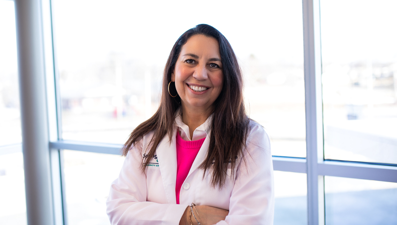 Female with long dark hair wearing white medical coat smiling in front of windows