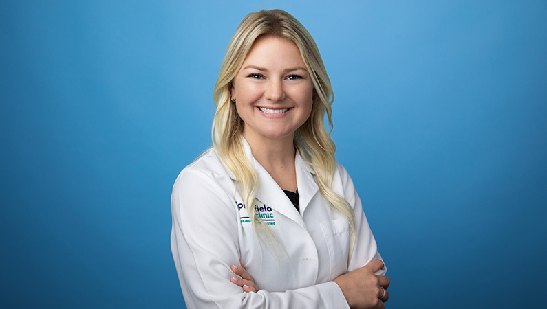 Female physician assistant wearing white medical coat smiling in front of light blue backdrop.