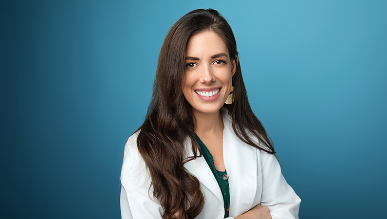 Female nurse with long brown hair wearing white medical coat smiling in front of blue photo backdrop.