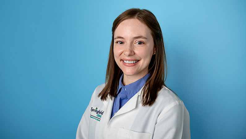 Female physician assistant in white medical coat smiling in front of light blue photo backdrop.
