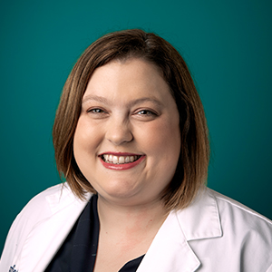 Female nurse practitioner in white coat smiling in front of a teal blue background.