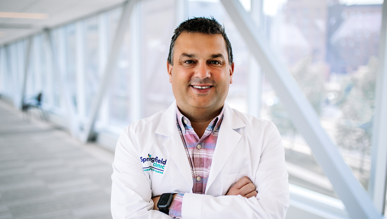 Male wearing white medical coat with arms crossed smiling in well-lit walkway