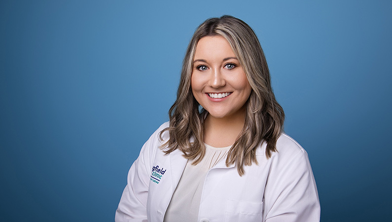 Female nurse practitioner wearing white medical coat smiling in front of blue photo backdrop