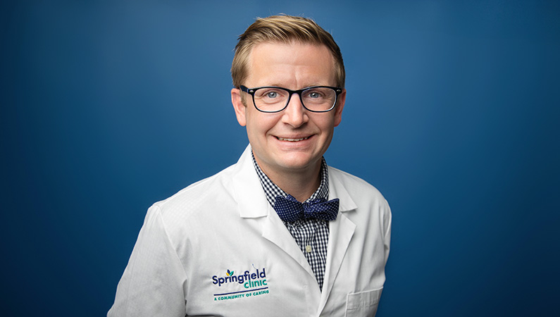 Cardiologist wearing white medical coat smiling in front of a light blue photo backdrop.